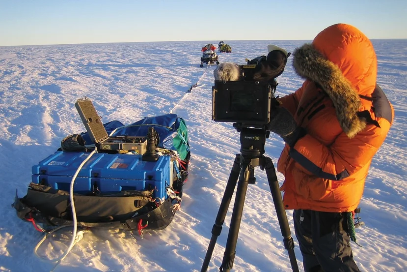 Polar researcher working a large-format camera on ice, gear packed in a blue Spacecase on a sled