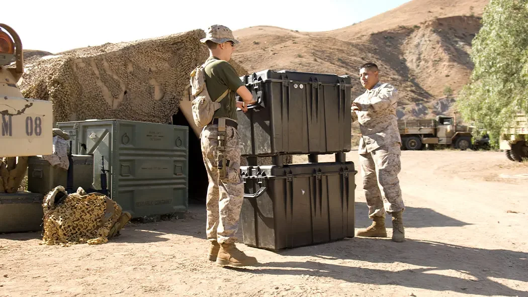 Two soldiers stacking large black Pelican cases at a desert field camp