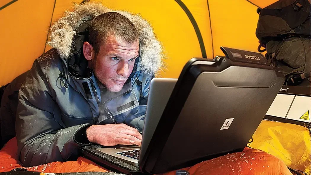 Expedition member using a laptop in a tent, the screen shielded by a Pelican laptop case