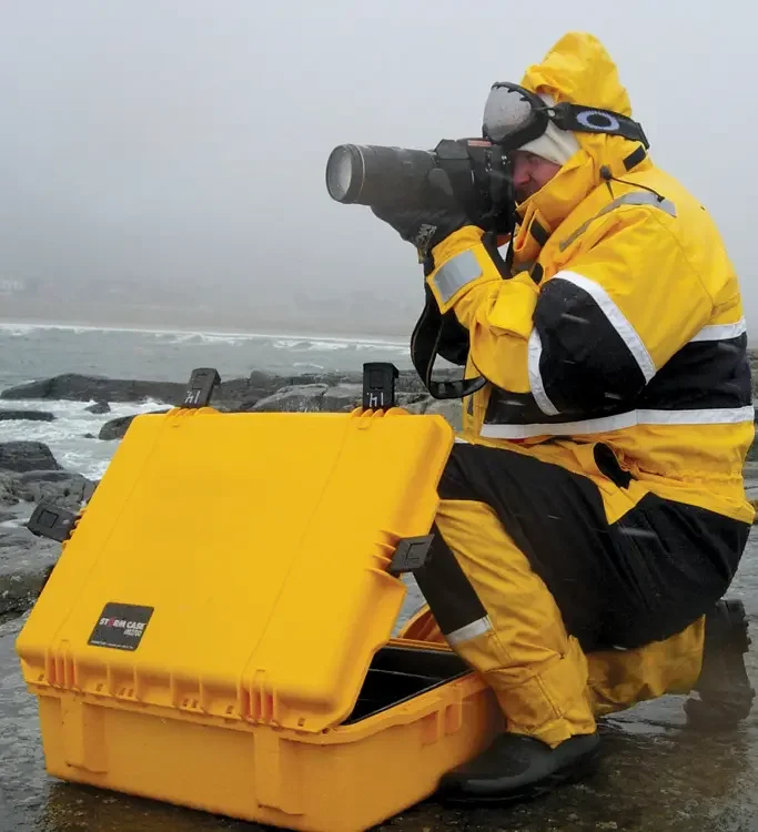 Photographer in wet weather gear on a rocky shore beside an open yellow Storm case