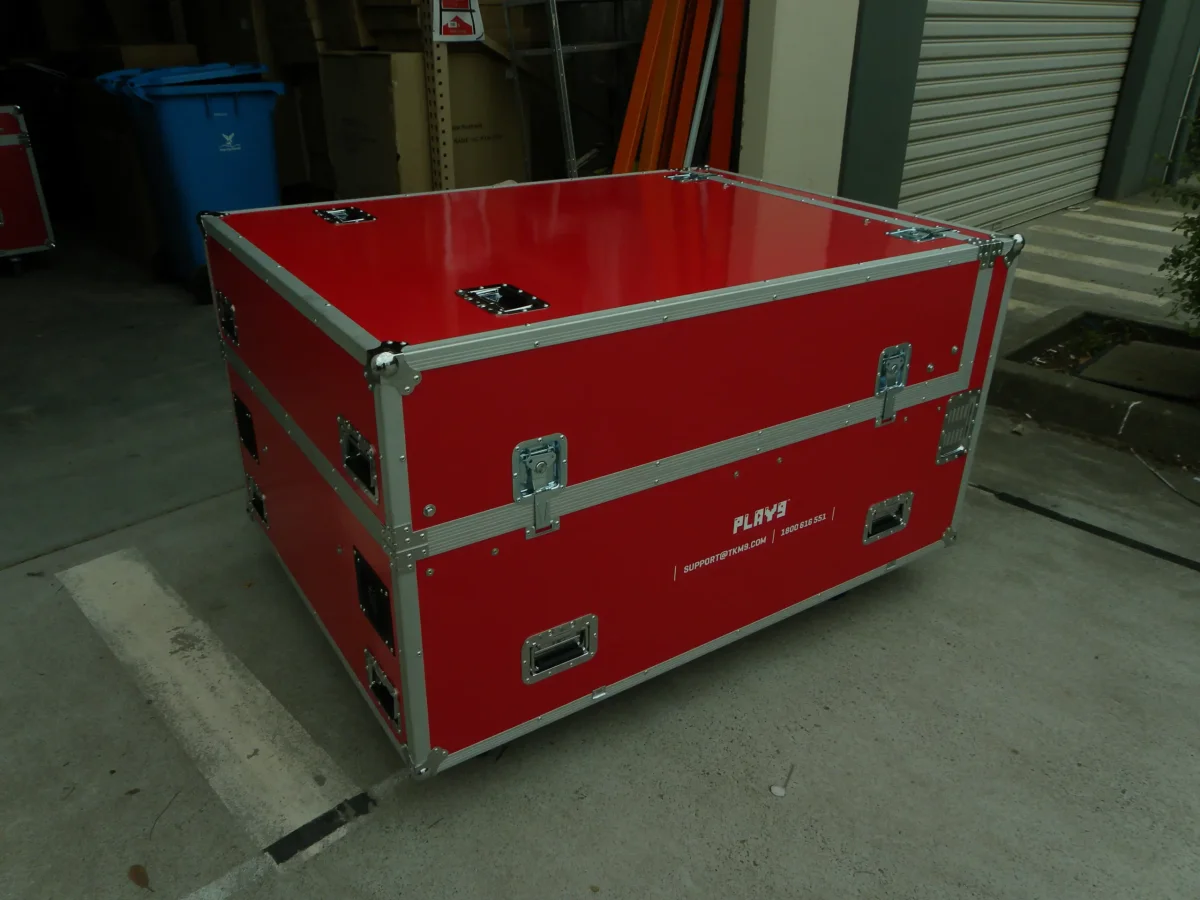 Red road case with a lift-off lid and recessed handles, staged at the roller door